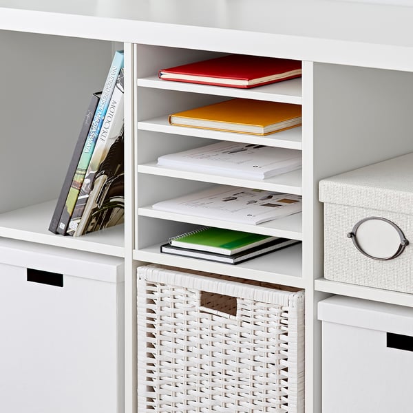 White KALLAX shelf unit with four vertical dividers, holding colorful books and a wicker basket.
