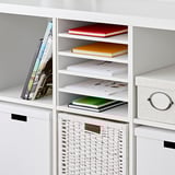 White KALLAX shelf unit with four vertical dividers, holding colorful books and a wicker basket.