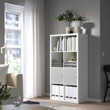 White KALLAX shelving unit, gray walls, light-filled room, books, plants, and minimalist decor. Tall, corner-placed, modern storage.