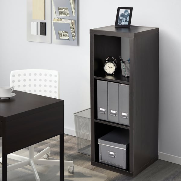 Black KALLAX bookshelf stands near a desk, holding clock, boxes, and photo. The shelves are modular, storing items neatly.