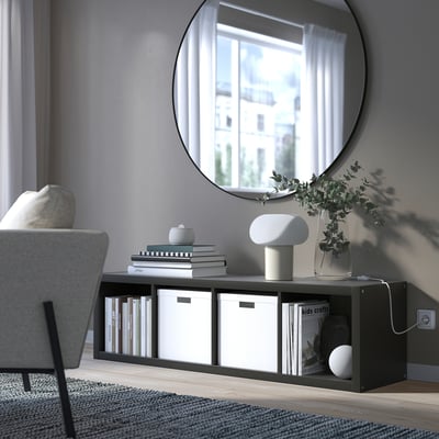 KALLAX shelving unit in grey with white boxes, lamp, and plant near a large window and mirror, reflecting a building.