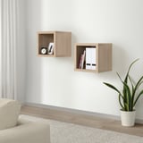 KALLAX wall shelves, light wood. Small, square cubes hold books and clock. Beside a beige chair and plant.