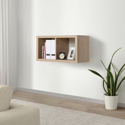 Floating KALLAX shelf in light brown holding books, clock, and framed photo. Next to green plant, light-colored couch, and gray rug.