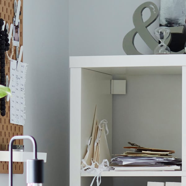 White KALLAX shelving unit against wall, stacked with papers and books. Includes a pegboard with notes and decorations.