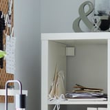 White KALLAX shelving unit against wall, stacked with papers and books. Includes a pegboard with notes and decorations.