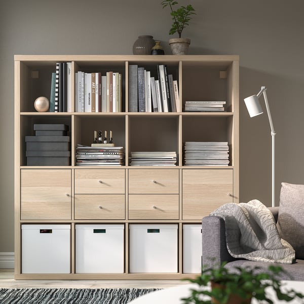 Wooden KALLAX shelf with books and decor, drawers, gray couch, white lamp.