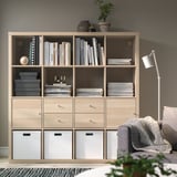 Wooden KALLAX shelf with books and decor, drawers, gray couch, white lamp.