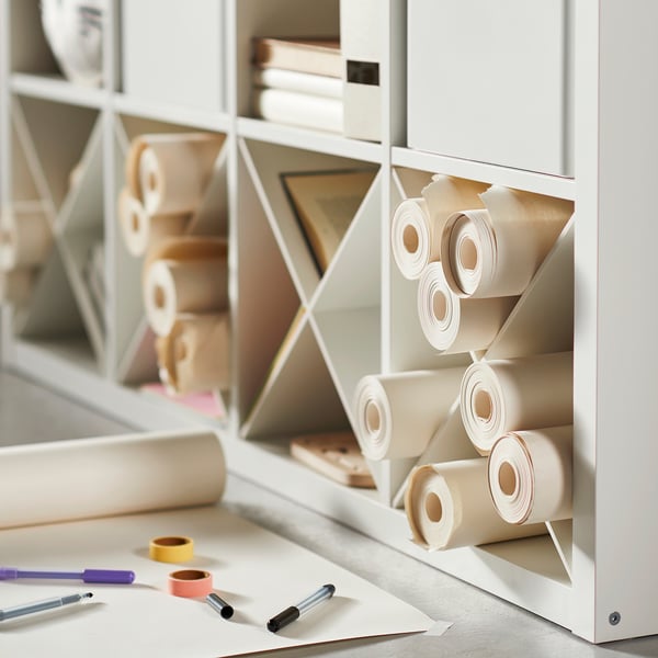White KALLAX shelving unit with compartments holding rolls of beige paper. On table, colorful pens and tape.