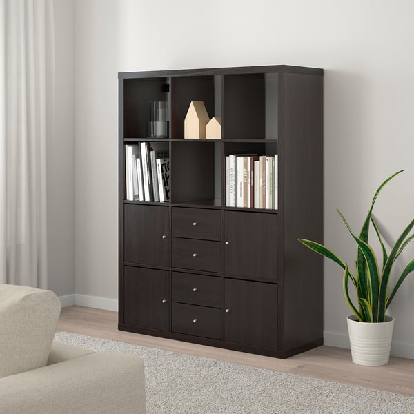 Black KALLAX bookshelf with drawers and shelves, storing books, vases, and decor next to a beige couch and plant.