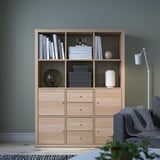 KALLAX light oak bookcase with drawers, hosting books, decorative items, and a lamp, fits well in a cozy green room with a gray couch nearby.
