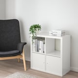 White ikea KALLAX shelving unit with books and plant, next to large grey chair on light wooden floor.