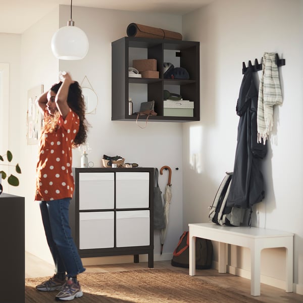 Woman dances in entryway beside black KALLAX storage unit. Bright light fixture above; white bench and hook rack to right.