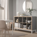 KALLAX light gray sideboard with wooden accents, holding vases, bowls, and plants, against a light gray wall beneath a round mirror.