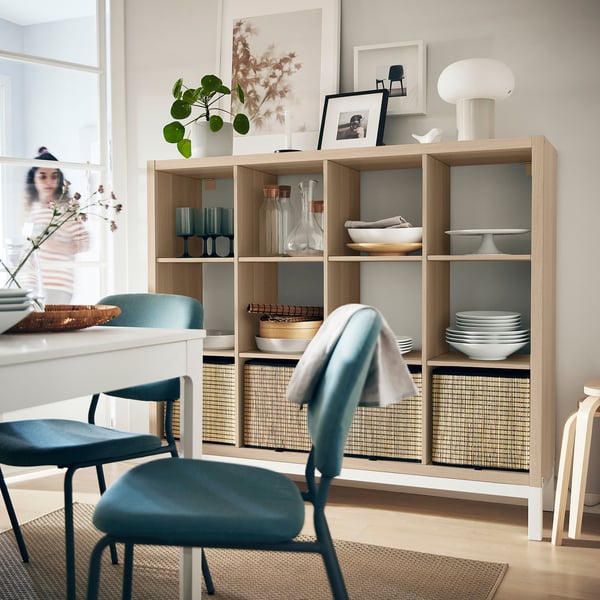 Cozy dining room with KALLAX shelves holding dishes, vases, books, and plants. Blue chair, white lamp, bright atmosphere.