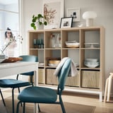 Cozy dining room with KALLAX shelves holding dishes, vases, books, and plants. Blue chair, white lamp, bright atmosphere.