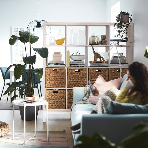 Woman on blue sofa using phone, KALLAX shelving with plants, bright room.