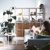 Woman on blue sofa using phone, KALLAX shelving with plants, bright room.