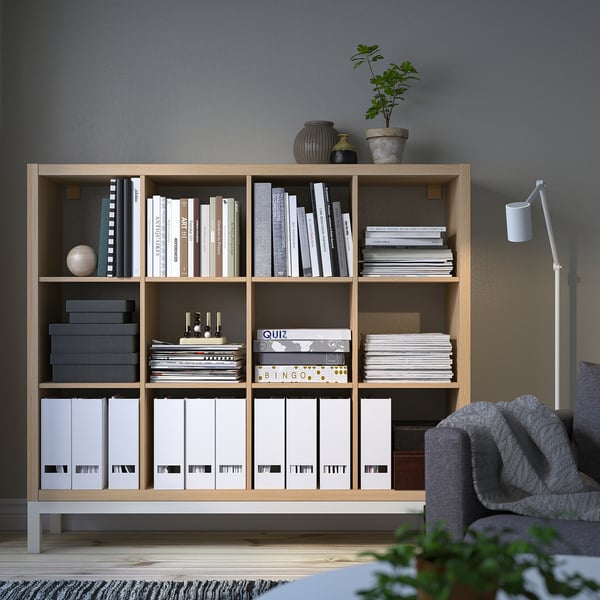 KALLAX shelf holds books, games, files. Tall with multiple compartments. Grey chair, white lamp, green plant nearby.