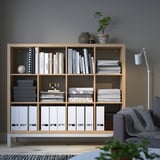 KALLAX shelf holds books, games, files. Tall with multiple compartments. Grey chair, white lamp, green plant nearby.