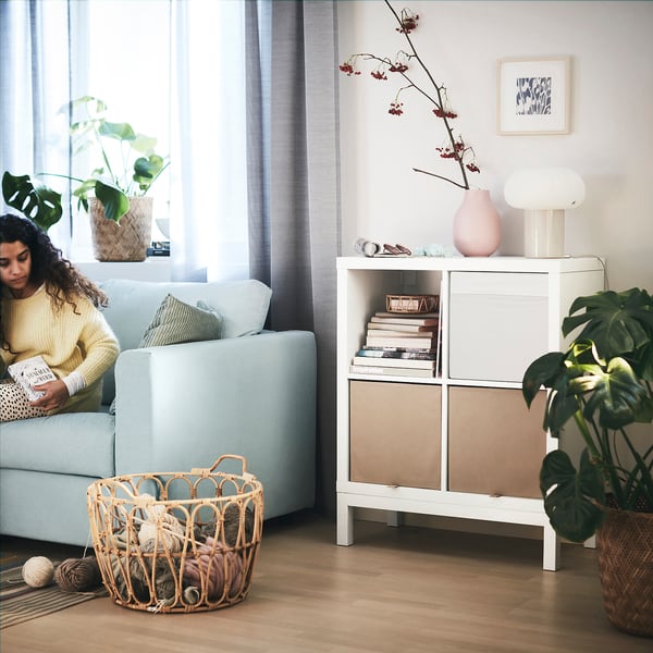 Woman reads on light blue sofa by KALLAX shelf with yarn basket, books, and plant.