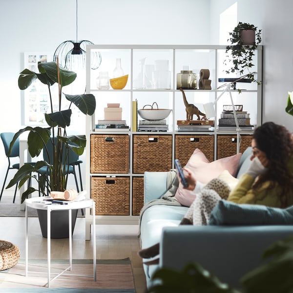 Modern living room with KALLAX shelves, blue sofa, pink pillows, plant, person on phone.
