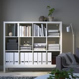 White KALLAX shelf with items, between armchair and lamp, topped with plants.