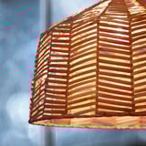 Hand-woven lamp shade, intricate herringbone pattern, warm glowing light. Crafted from rattan, emits cozy, inviting ambiance.