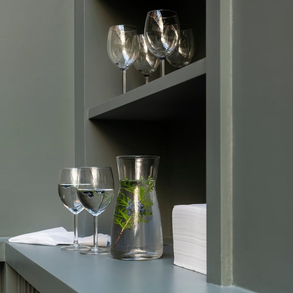 Clear glass carafe with greenery, four wine glasses on shelves. Gray shelves, minimalist decor.