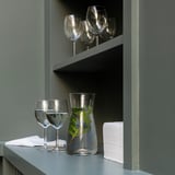 Clear glass carafe with greenery, four wine glasses on shelves. Gray shelves, minimalist decor.