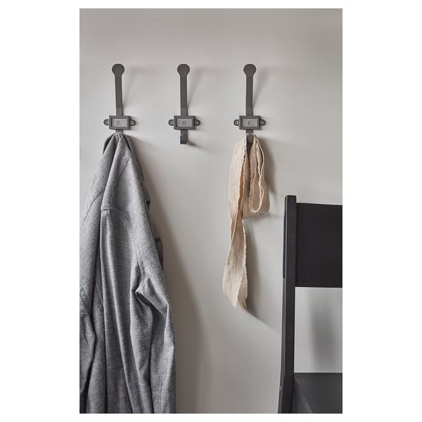 Three gray metal hooks on a wall hang a gray coat, beige scarf, and brown scarf. Modern design with square labels for personalized use.