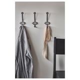 Three gray metal hooks on a wall hang a gray coat, beige scarf, and brown scarf. Modern design with square labels for personalized use.