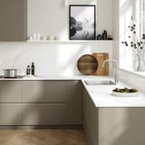 Modern kitchen with white quartz countertop, wooden board, fruits, grey cabinets, artwork, and window with dried flowers.