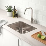 Kitchen sink with faucet, knives, limes, countertop with bottles, plant, and cutting board nearby.