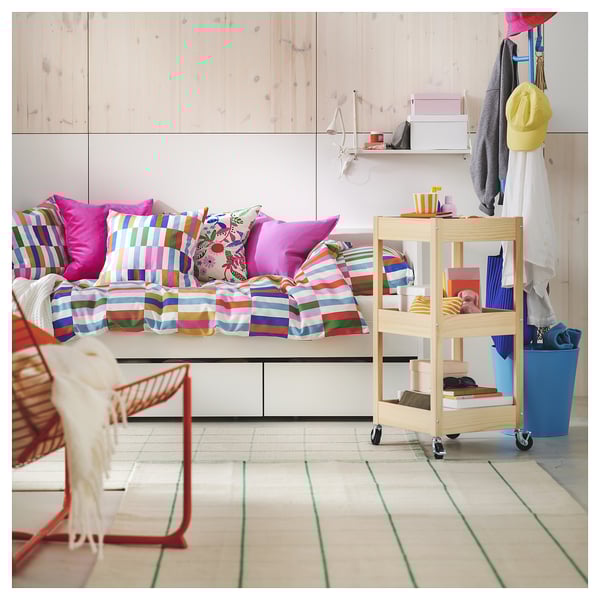 Colorful bedroom with striped bedding, pillows, and cart items. White walls, wooden floors, pastel organizers.