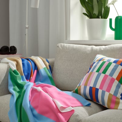 Bright, multicolored KLIBBKORSÖRT throw blanket on large beige couch, next to green plant and colorful pillow, providing vibrant accent.