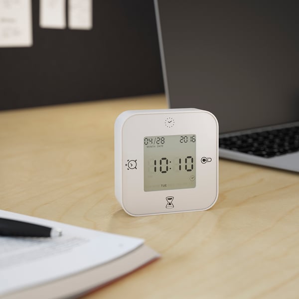 A square alarm clock displays 10:10 on a wooden desk. It shows date and time, one of its four functions.