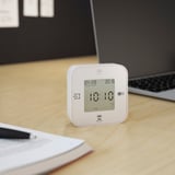 A square alarm clock displays 10:10 on a wooden desk. It shows date and time, one of its four functions.