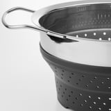 Collapsible stainless steel colander with silicone strainer, resting on edge, black and silver.