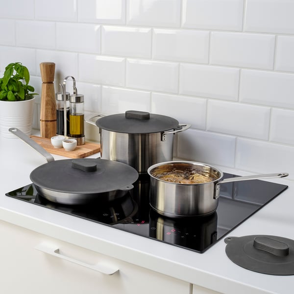 Three stainless steel pots with black silicone lids are on a modern stove, indicating they can be used on hobs and in ovens.