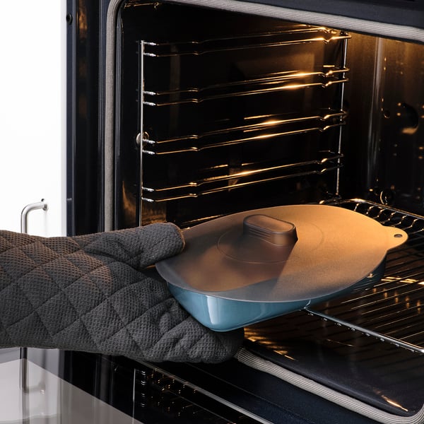 Person placing a blue dish with a round brown silicone lid into an oven. The lid helps keep food fresh and can be used in various kitchen settings.