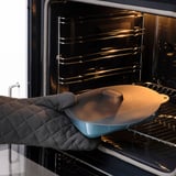 Person placing a blue dish with a round brown silicone lid into an oven. The lid helps keep food fresh and can be used in various kitchen settings.
