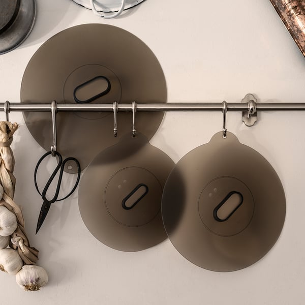 Three brown silicone lids hang from a metal bar with hooks, designed for various pots and pans, featuring a vacuum seal and versatile kitchen use.