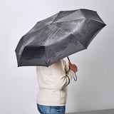 A person holding an open black KNALLA umbrella, providing rain protection. The umbrella is foldable and has a handle with a carrying strap.