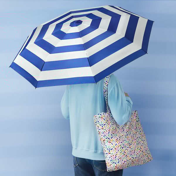 A person holds a blue and white striped umbrella, standing against a blue background, with a colorful tote bag on their shoulder.
