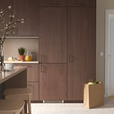 Modern kitchen with dark wood cabinets, tall KÖLDGRADER fridge, countertop with plants, jars, bread, and groceries on floor, small bar table.