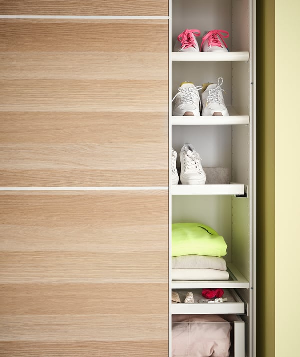 Wooden closet with white shelves holding green shirt, white and gray sneakers, pink sandals, and white socks.