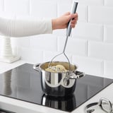 Person is using a KONCIS potato masher on a stove. The masher has a long handle for easy reach into pots, creating smooth vegetable mash.