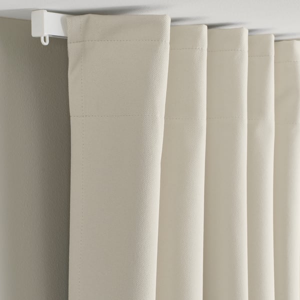 Beige KORGMOTT block-out curtains hang with pleated folds, mounted by a white curtain rod. They block light and offer privacy.