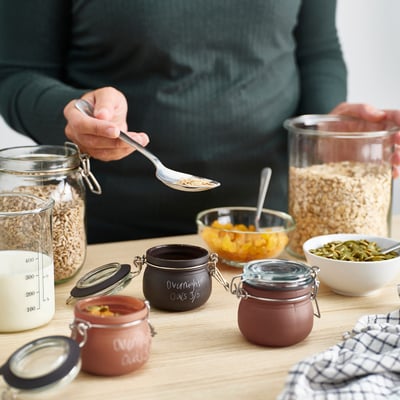 Person preparing overnight oats using KORKEN jars, with ingredients and bowls on a wooden table.