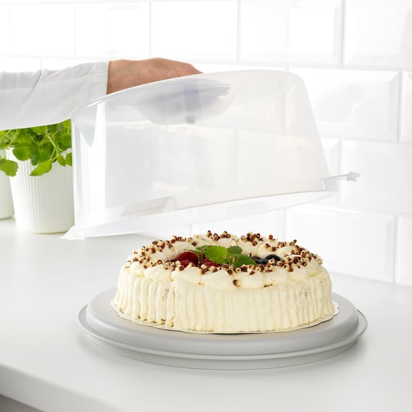 A cake in a carrier with a clear lid, placed on a countertop. The carrier has grooves and smooth sides for different pastries.